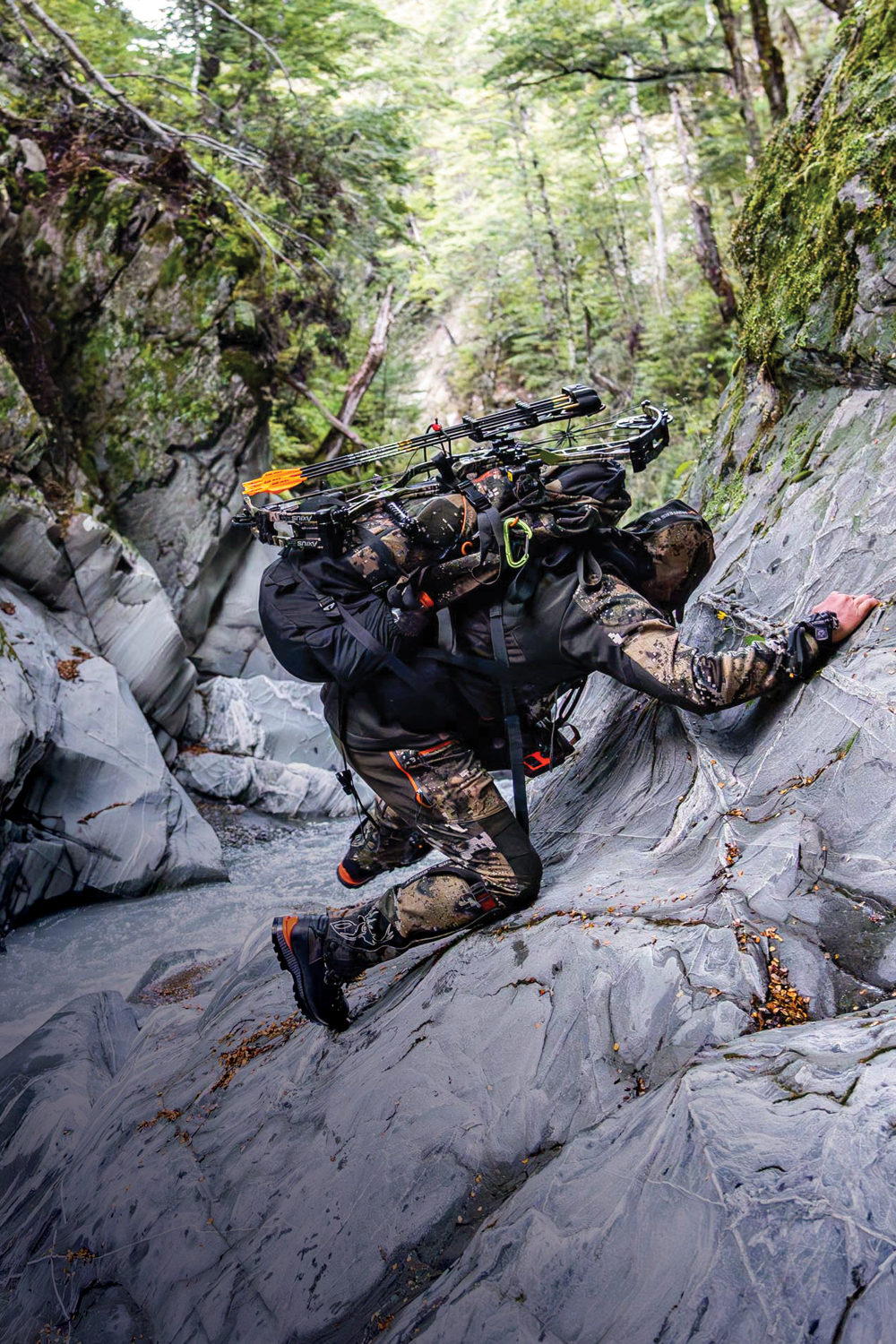 Person in camouflage gear climbing a rocky mountain path with hunting equipment.