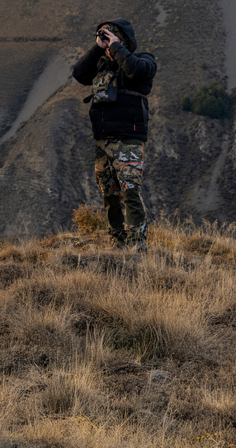 Person in camouflage clothing and a hooded jacket standing on a grassy hillside, looking through binoculars.