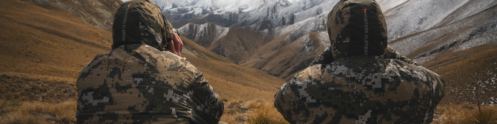 Two people in camouflage gear looking out at a mountainous landscape with snow-capped peaks.