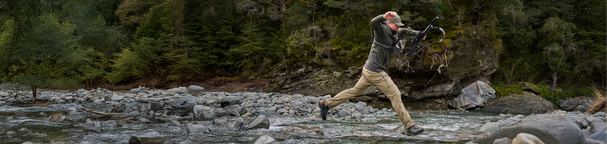Spur Pants homepage desktop banner showing Cody jumping over a river wearing the Tussock Spur Pants.