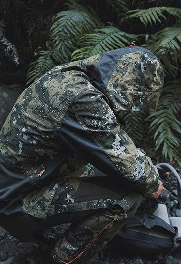 Person in camouflage jacket and pants crouching in a forest setting